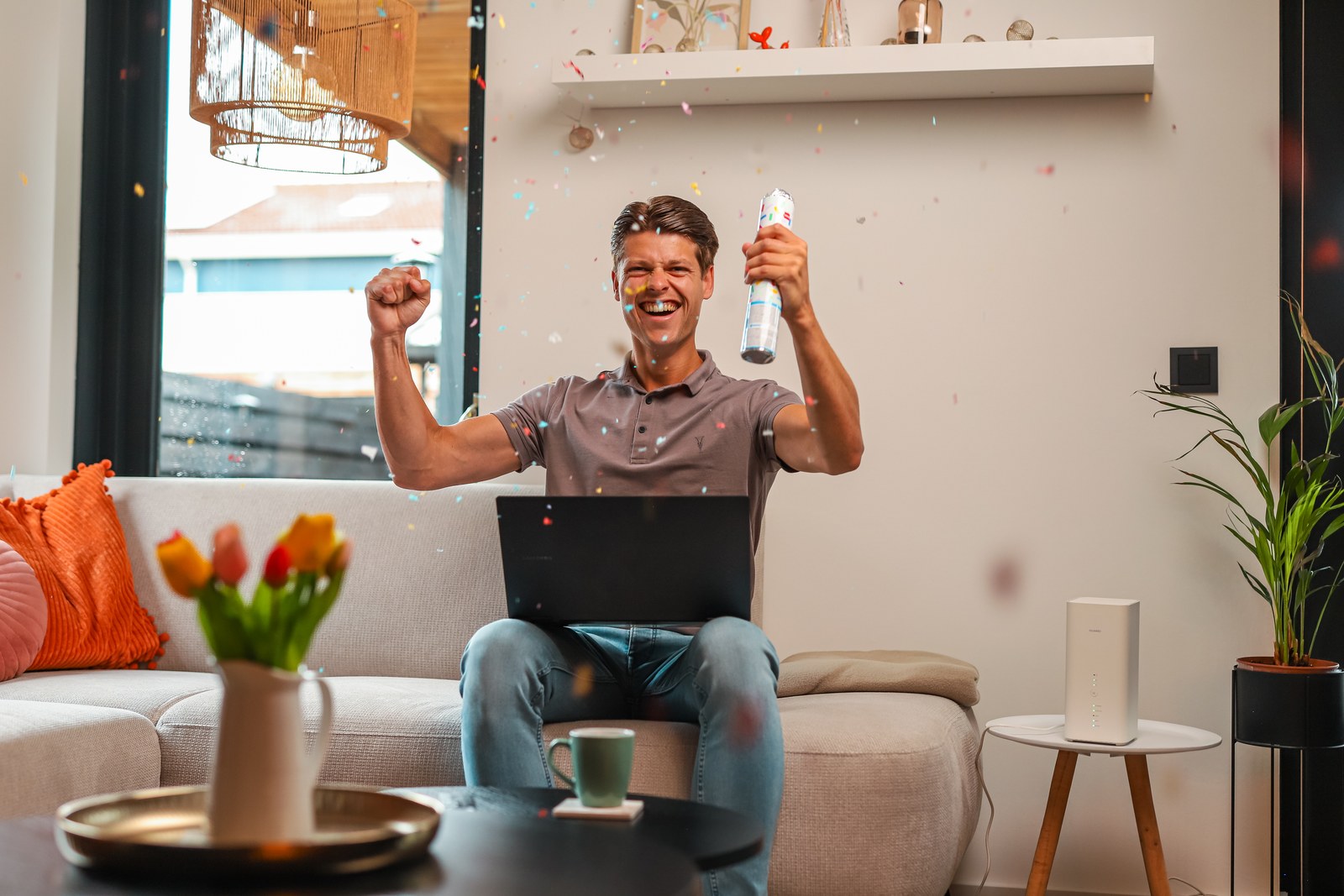 Man juicht met een laptop op schoot op de bank, met het 5G-modem op de bijzettafel