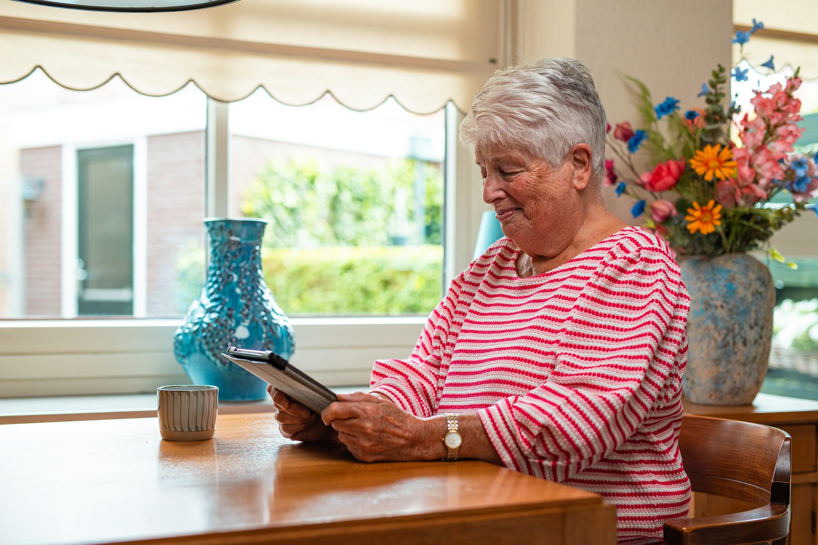 Vrouw gebruikt een tablet aan tafel bij het raam, met bloemen op de achtergrond