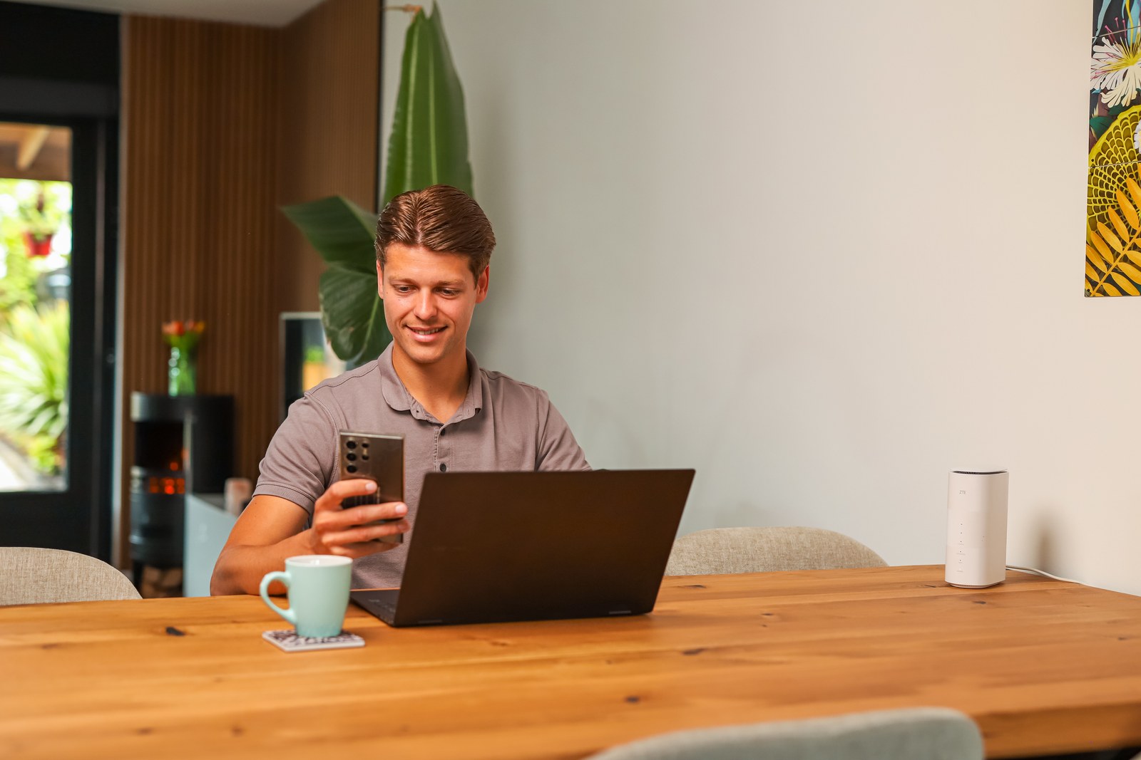 Man aan een bureau kijkt op zijn telefoon, met een laptop en het modem voor hem